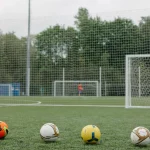 Empat bola sepak berjajar di lapangan rumput dengan gawang latihan di belakangnya