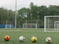 Empat bola sepak berjajar di lapangan rumput dengan gawang latihan di belakangnya