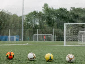 Empat bola sepak berjajar di lapangan rumput dengan gawang latihan di belakangnya