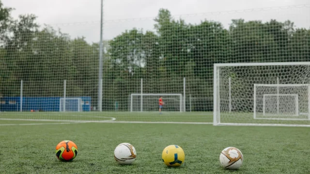 Empat bola sepak berjajar di lapangan rumput dengan gawang latihan di belakangnya