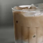 Gelas bening berisi kopi dingin berlapis susu dengan es batu dan buih di permukaannya