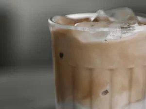 Gelas bening berisi kopi dingin berlapis susu dengan es batu dan buih di permukaannya