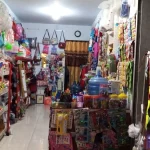 Deretan rak warung kelontong penuh tas, mainan, galon air, dan barang kebutuhan harian