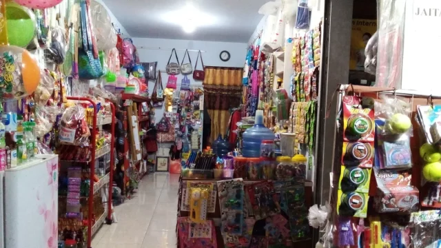 Deretan rak warung kelontong penuh tas, mainan, galon air, dan barang kebutuhan harian