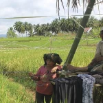 Ilustrasi dua orang duduk di dangau bambu sambil memegang tali penghalau burung di tengah hamparan sawah