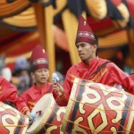 Pemain gandang Minangkabau berbaju merah dan bertopi tinggi menabuh gendang bermotif