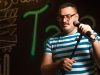Ilustrasi seorang pelawak tunggal berbicara ke mikrofon di panggung berlatar lampu neon.