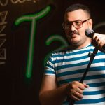 Ilustrasi seorang pelawak tunggal berbicara ke mikrofon di panggung berlatar lampu neon.