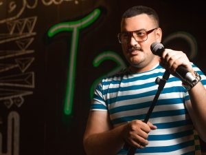 Ilustrasi seorang pelawak tunggal berbicara ke mikrofon di panggung berlatar lampu neon.