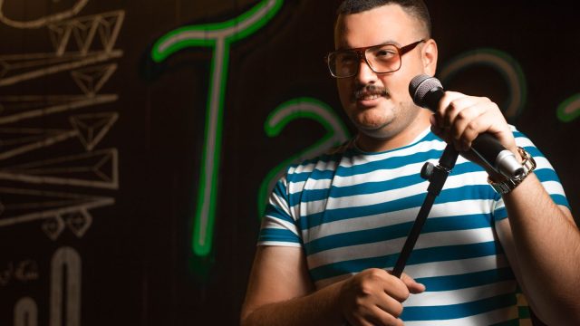 Ilustrasi seorang pelawak tunggal berbicara ke mikrofon di panggung berlatar lampu neon.