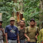 Sekelompok laki-laki berdiri di dalam hutan sambil menggendong kayu dan keranjang anyaman