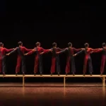 Ilustrasi delapan penari berkostum merah dan biru tua berdiri berjajar di atas bangku panjang