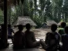 Ilustrasi warga kampung duduk melingkar di beranda kayu menghadap rumah beratap daun sagu