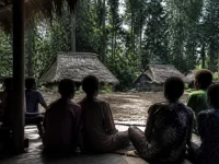 Ilustrasi warga kampung duduk melingkar di beranda kayu menghadap rumah beratap daun sagu