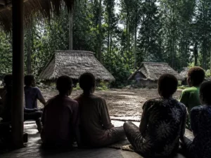 Ilustrasi warga kampung duduk melingkar di beranda kayu menghadap rumah beratap daun sagu