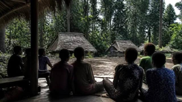 Ilustrasi warga kampung duduk melingkar di beranda kayu menghadap rumah beratap daun sagu