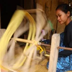 Ilustrasi perempuan memintal benang dengan roda pintal tradisional di Banten