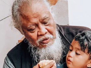 Kakek berjanggut putih memangku cucu laki-lakinya sambil menyuapkan kue di teras rumah