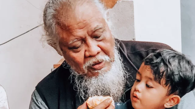Kakek berjanggut putih memangku cucu laki-lakinya sambil menyuapkan kue di teras rumah