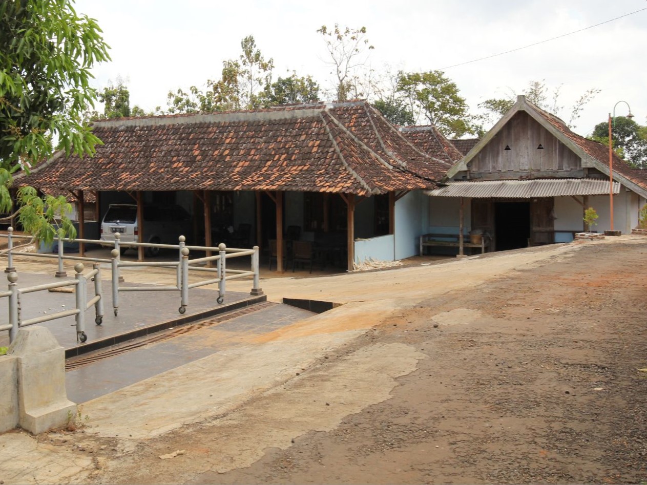 Sebuah rumah tradisional Jawa berdinding kayu dengan atap genting memanjang berdiri di tepi jalan tanah.