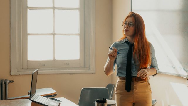 Ilustrasi seorang perempuan berbicara di depan kelas sambil memegang catatan.