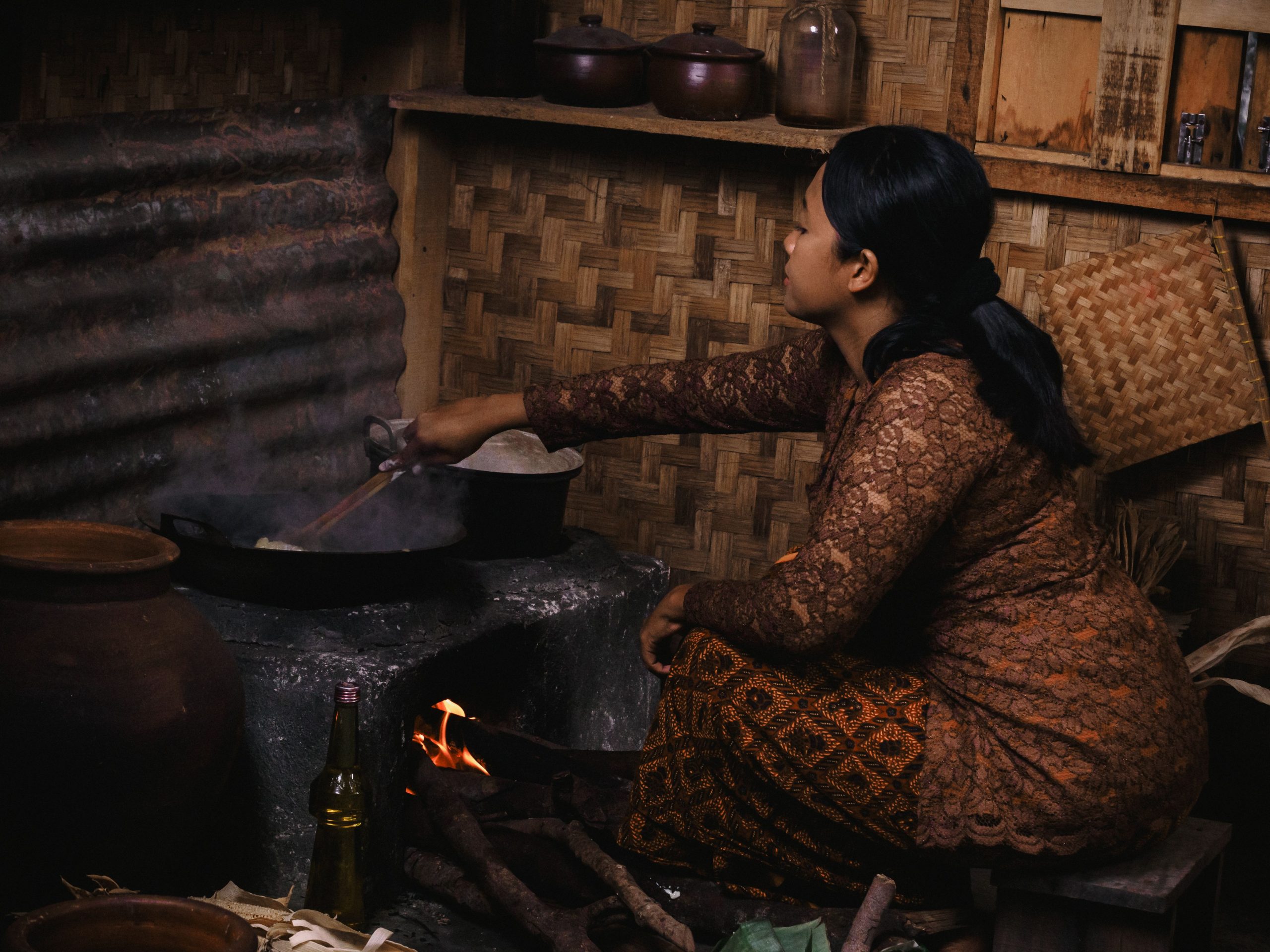 Ilustrasi seorang perempuan memasak di tungku kayu dalam dapur tradisional.