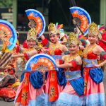 Ilustrasi sekelompok penari berkostum warna-warni tampil dalam pertunjukan tradisional di ruang terbuka.
