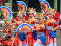 Ilustrasi sekelompok penari berkostum warna-warni tampil dalam pertunjukan tradisional di ruang terbuka.