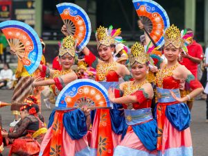 Ilustrasi sekelompok penari berkostum warna-warni tampil dalam pertunjukan tradisional di ruang terbuka.
