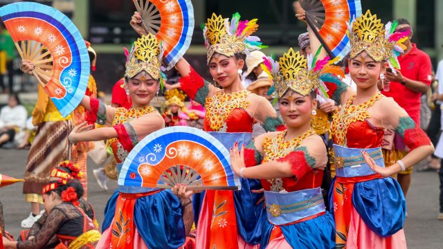 Ilustrasi sekelompok penari berkostum warna-warni tampil dalam pertunjukan tradisional di ruang terbuka.