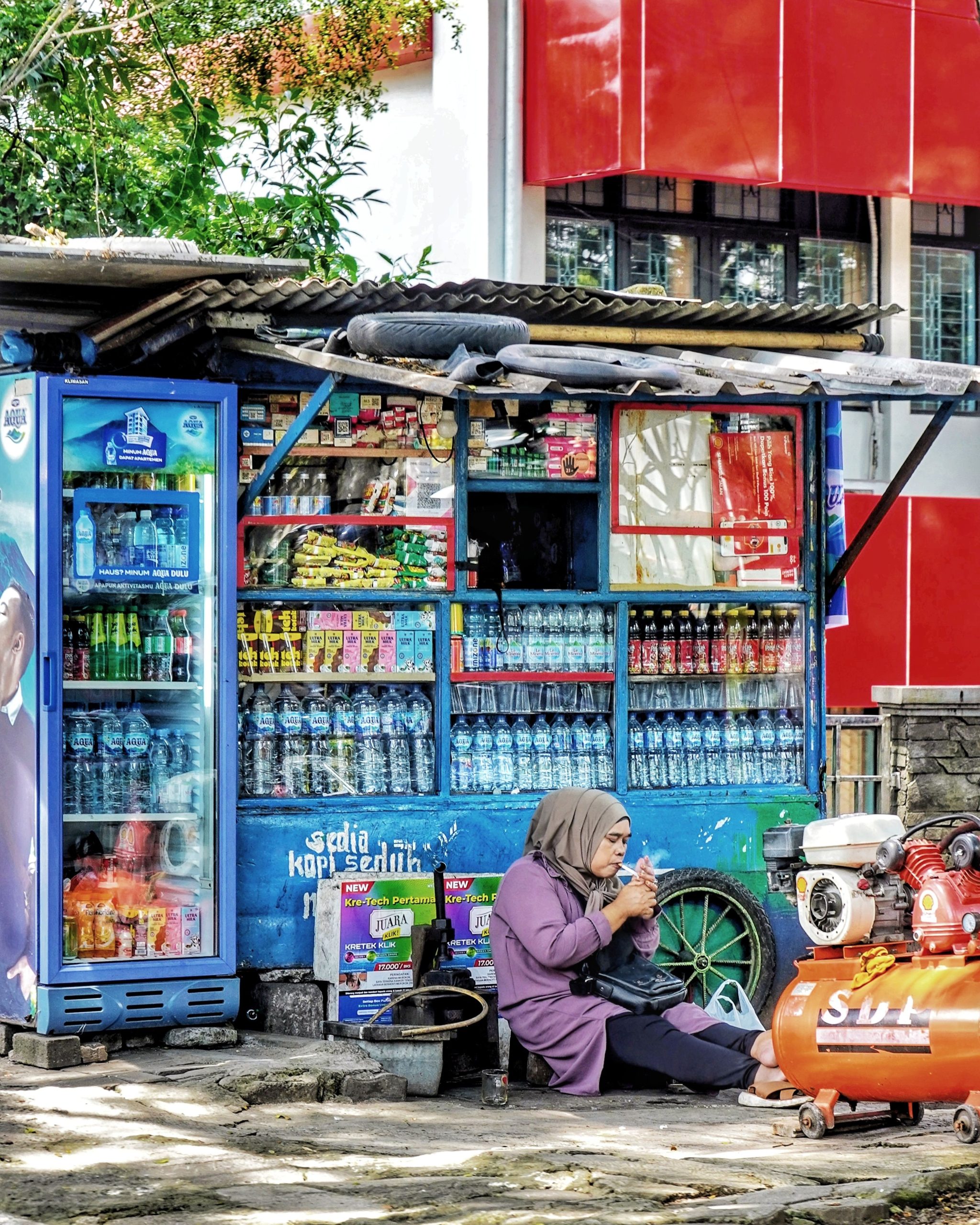 Ilustrasi warung kecil di tepi jalan dengan aneka dagangan tergantung dan pemiliknya duduk di depan.