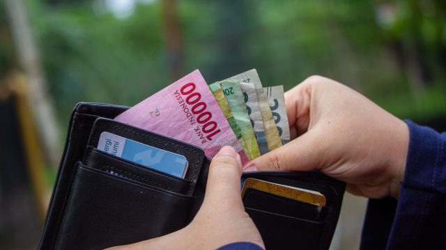 Ilustrasi tangan membuka dompet berisi lembaran uang rupiah dan kartu.