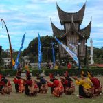 Para pemain randai duduk melingkar di halaman berumput di depan bangunan beratap gonjong saat pertunjukan berlangsung.