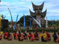 Para pemain randai duduk melingkar di halaman berumput di depan bangunan beratap gonjong saat pertunjukan berlangsung.