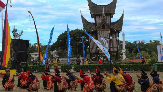 Para pemain randai duduk melingkar di halaman berumput di depan bangunan beratap gonjong saat pertunjukan berlangsung.