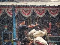 Ilustrasi seorang pengendara sepeda motor menembus hujan deras di jalan kampung.