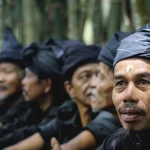 Sejumlah laki-laki Kajang berpakaian dan berikat kepala hitam duduk berjajar di depan rumpun bambu