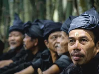 Sejumlah laki-laki Kajang berpakaian dan berikat kepala hitam duduk berjajar di depan rumpun bambu
