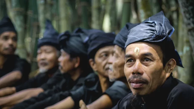 Sejumlah laki-laki Kajang berpakaian dan berikat kepala hitam duduk berjajar di depan rumpun bambu