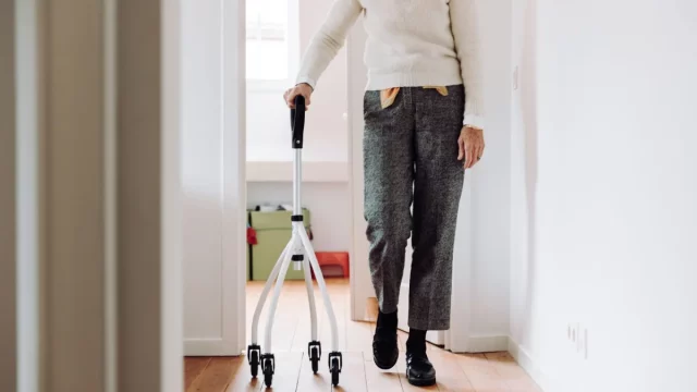 Seorang lanjut usia berjalan di dalam rumah menggunakan alat bantu rollator