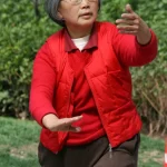 Perempuan lansia mengenakan rompi merah berlatih gerakan tai chi di taman terbuka