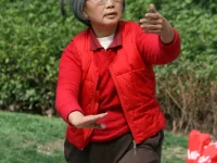 Perempuan lansia mengenakan rompi merah berlatih gerakan tai chi di taman terbuka