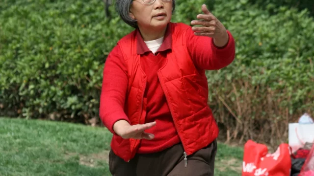 Perempuan lansia mengenakan rompi merah berlatih gerakan tai chi di taman terbuka