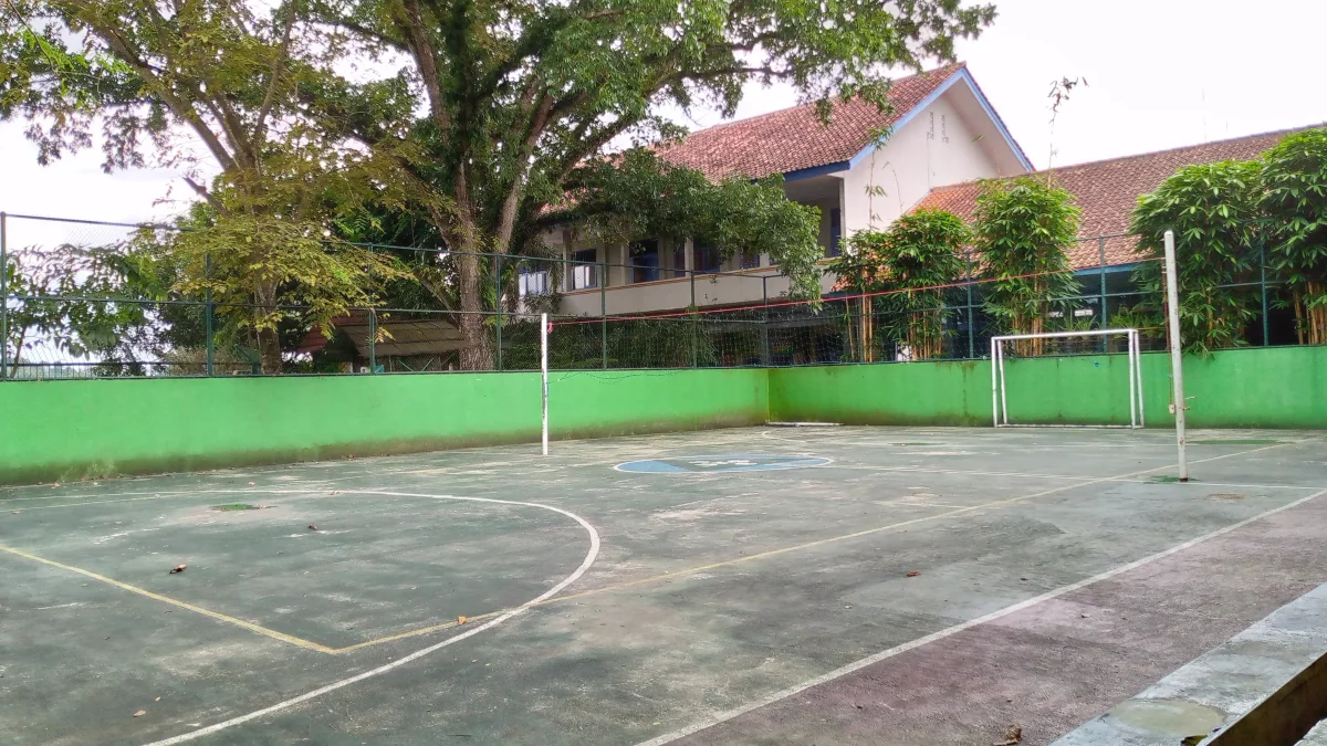 Lapangan olahraga berpagar hijau dengan gawang futsal dan tiang net voli di halaman sekolah