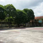 Lapangan futsal terbuka yang kosong dengan ring basket dikelilingi pepohonan dan gedung sekolah