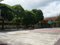 Lapangan futsal terbuka yang kosong dengan ring basket dikelilingi pepohonan dan gedung sekolah