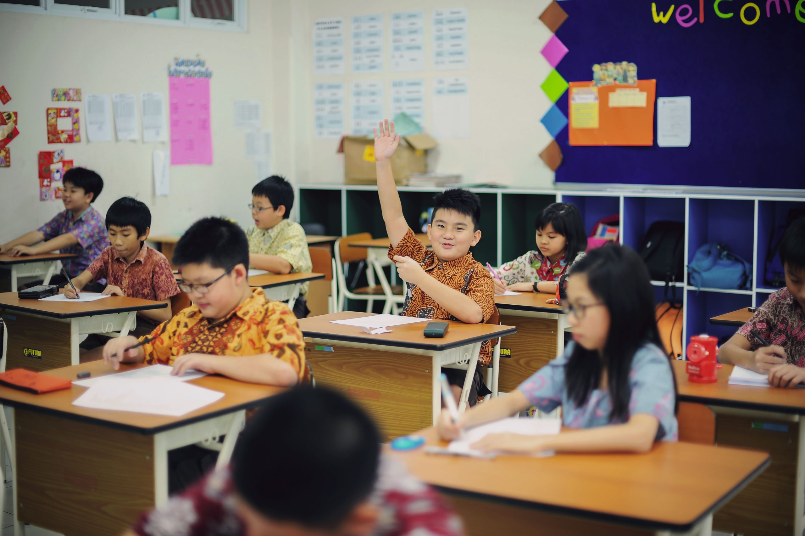 Ilustrasi murid-murid sekolah dasar mengerjakan tugas di meja kelas yang terang.