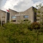 Gedung rektorat Universitas Andalas berdinding beton dengan tiang bendera Merah Putih, dikelilingi rimbun pepohonan.