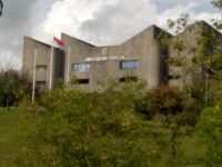 Gedung rektorat Universitas Andalas berdinding beton dengan tiang bendera Merah Putih, dikelilingi rimbun pepohonan.