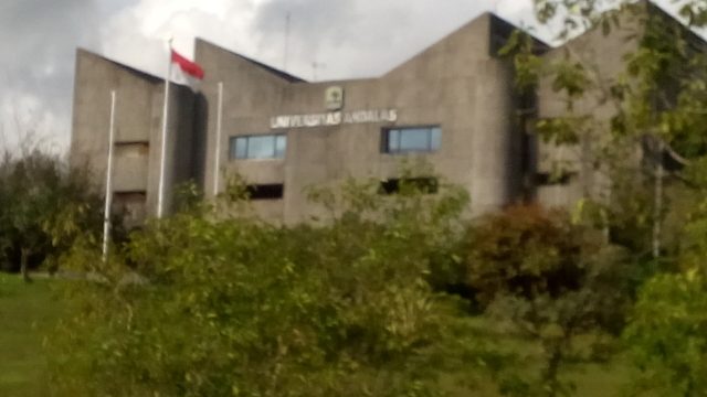 Gedung rektorat Universitas Andalas berdinding beton dengan tiang bendera Merah Putih, dikelilingi rimbun pepohonan.